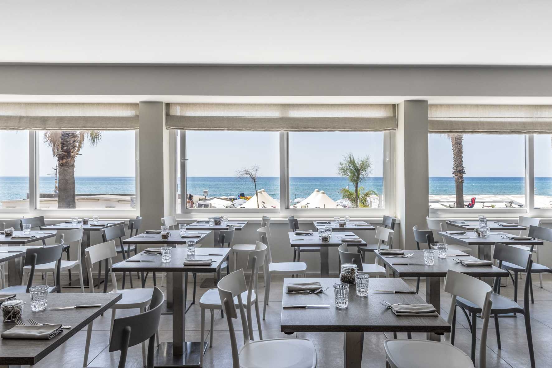 Sala apparecchiata, tavoli grigi e sedie bianche, tre finestroni sul mare e sulla spiaggia, Centosedici