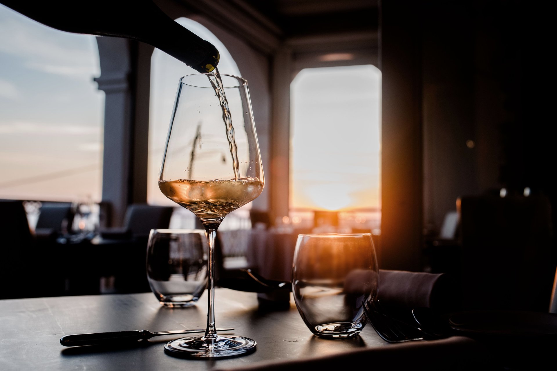 Vino bianco versato nel calice su tavolo scuro, controluce del tramonto dalle finestre ad arco, Il Granchio