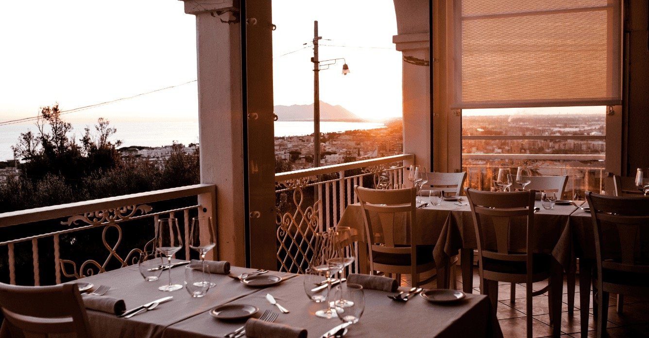 Sala in veranda apparecchiata, vetrate aperte sul panorama al tramonto: tetti di Terracina, mare, Circeo, Il Granchio