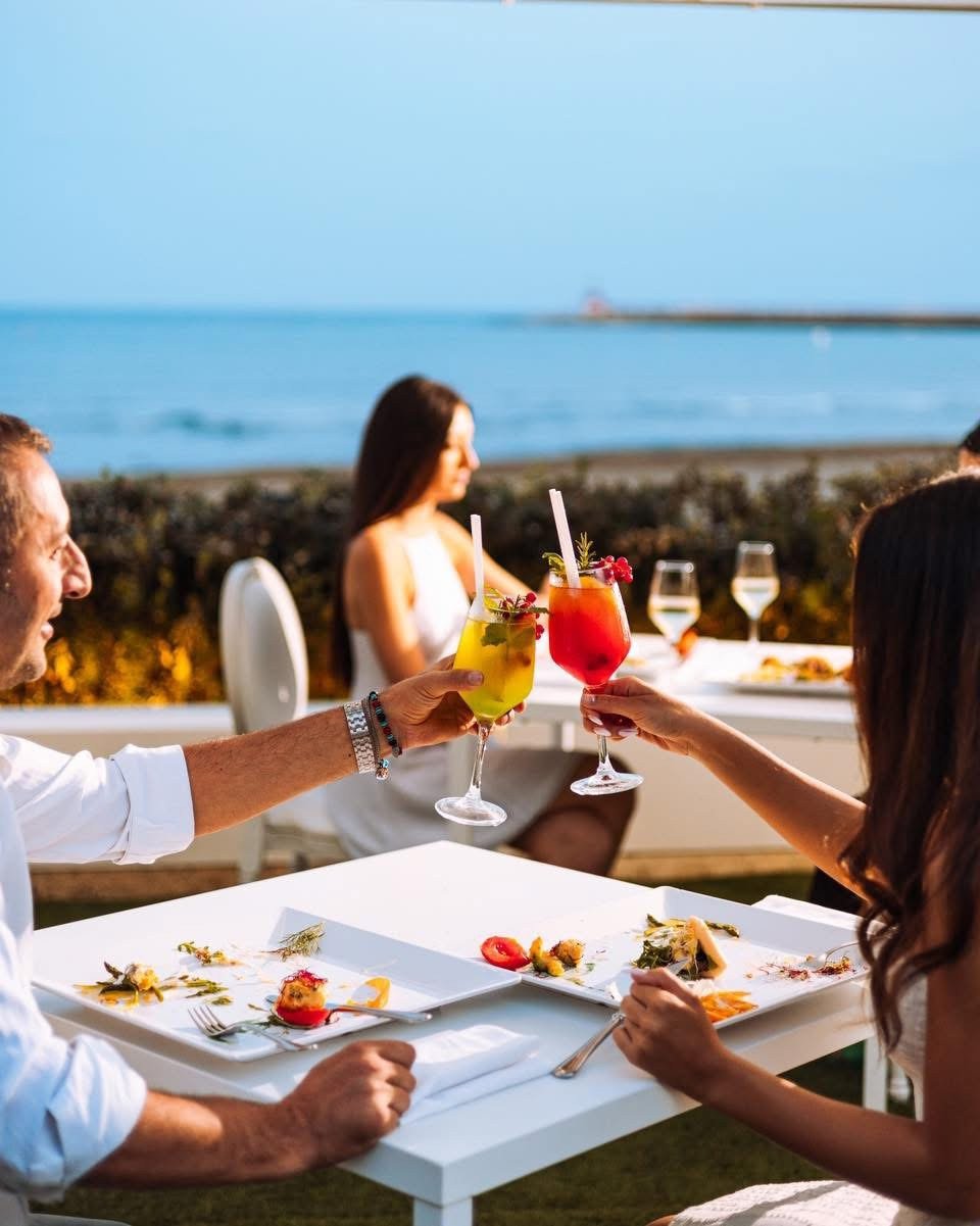 Coppia che brinda con due cocktail a un tavolo con piatti, dehors sul mare, molo col faro in fondo (stesso file di B-lepini-appia/foto/P6-terracina-la-capannina-brindisi.jpg), La Capannina