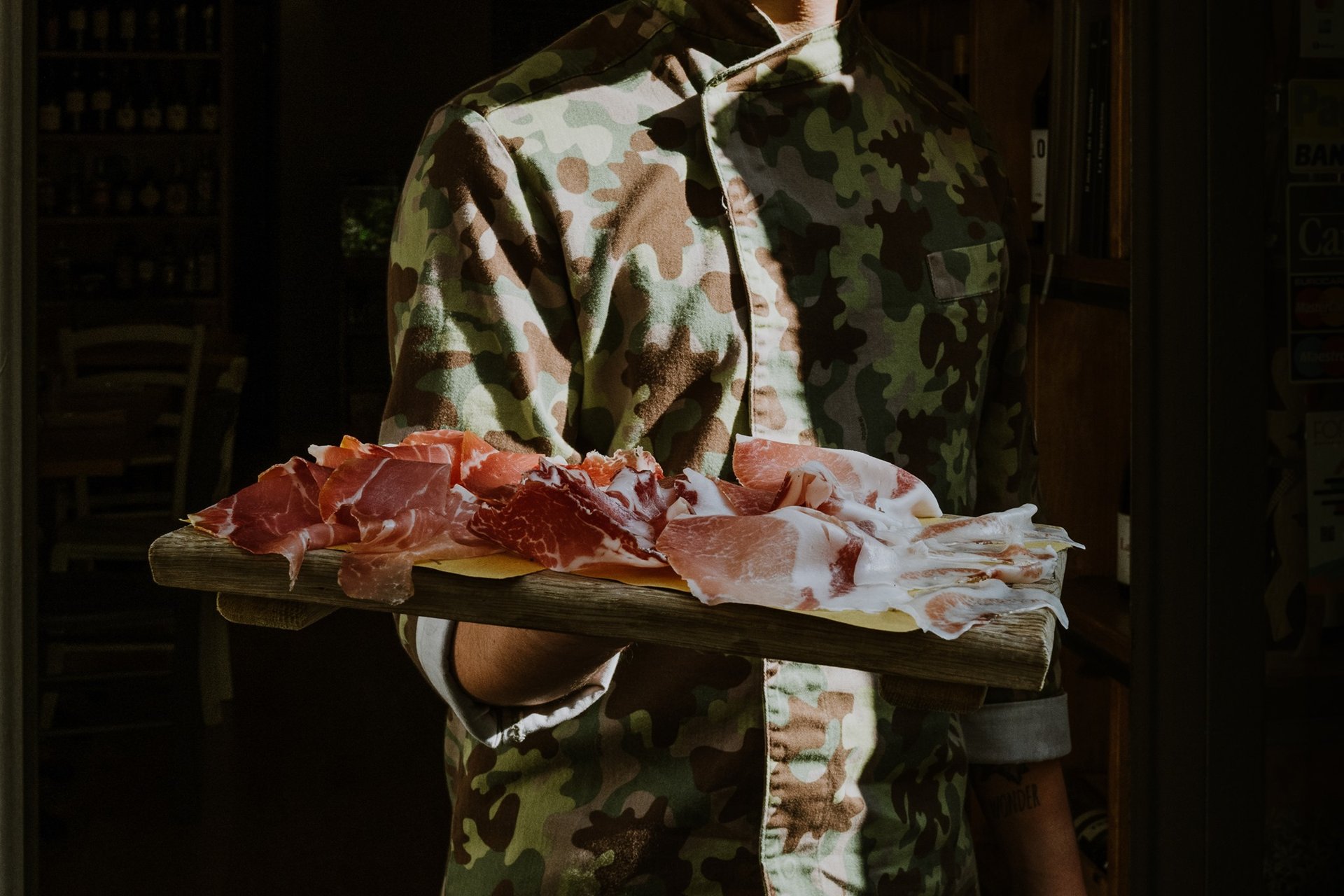 Persona in giacca da cuoco mimetica porta un tagliere di salumi sulla soglia del locale, controluce, Novecento