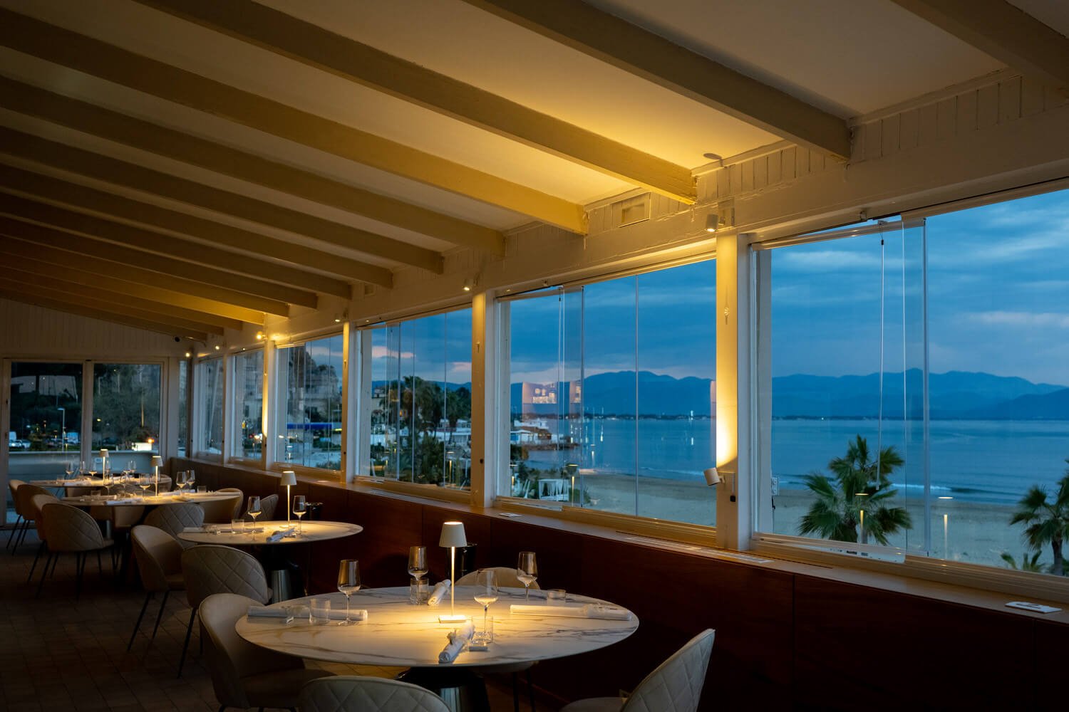 Sala all'imbrunire, tavoli rotondi con lampade accese, vetrate larghe sul mare e sulla costa, Ristorante Grappolo D’Uva Ricevimenti