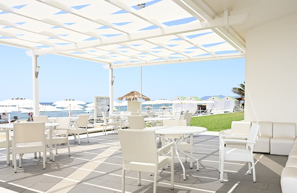 Terrazza coperta con tavoli e sedie bianche, ombrelloni del lido e mare dietro, Torre del Sole
